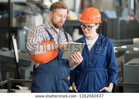 Young female engineer in uniform and helmet looking at tablet display while listening to colleague explanation of data Royalty-Free Stock Photo #1362060437
