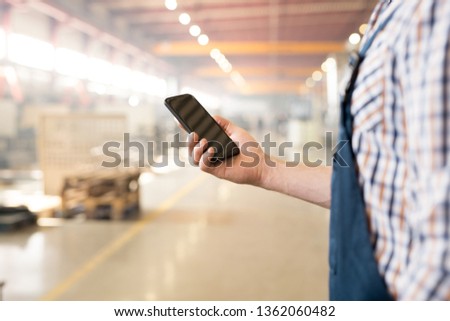Modern gadget with touchscreen held by young engineer or technician in workwear texting by workplace
