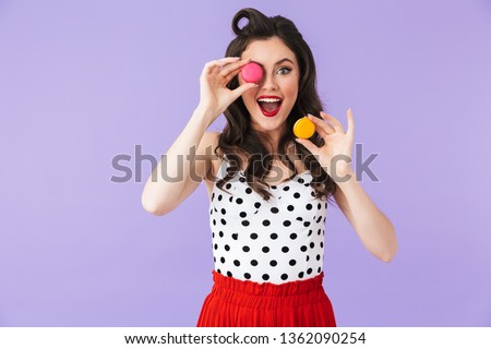 Photo of cheery pin-up woman 20s in vintage polka dot dress rejoicing while holding and eating macaron cookies isolated over violet background