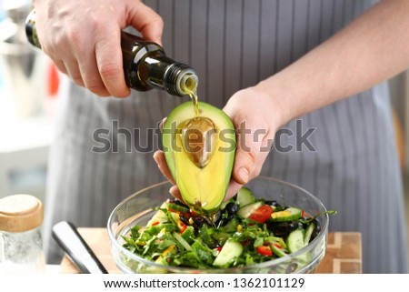 Male cook at home holds bottle of olive oil in his hand. Watering dressing avocado salad with bone seed incision concept on kitchen background closeup.