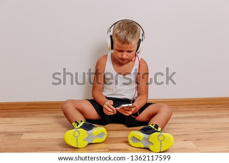 seven-year-old tanned child listening to music through large full-size headphones on a white background. A boy in bright green sneakers sits on the floor and uses a smartphone. Modern children
