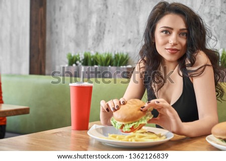 Pretty young female student having lunch in cafe. Brunette sitting at table, eating tasty hamburger, drinking sweet water looking at camera and smiling in pizzeria. Concept of fast food.