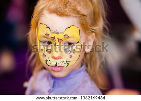 Child animator, artist's hand draws face painting to little girl. Child with funny face painting. Painter makes yellow leopard at girl's face. Children holiday, event, birthday party, entertainment.