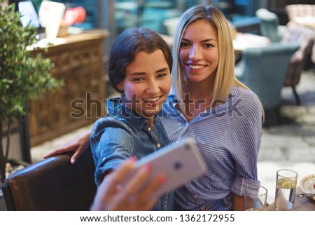 Two smiling young woman posing together for selfie on smartphone at table in restaurant