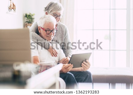couple of adult seniors smiling and looking at the tablet - retired people using technology at home - woman hug the man with love - forever together nice old people enjoy technology Royalty-Free Stock Photo #1362193283