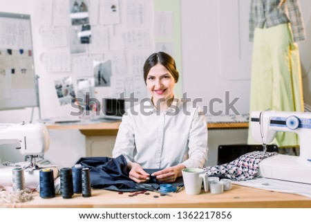 Smiling female fashion designer sitting at office desk. Dressmaker, tailor, works and seamstress concept - portrait of smiling fashion designer using sewing machine