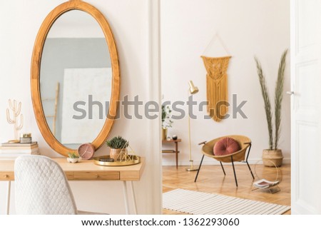 Stylish and cozy scanidnavian interiors of apartment with big mirror, dressing table, gold armchair, design accessories and furnitures, plant, palm leaves, yellow macrame. Modern home decor of rooms. 