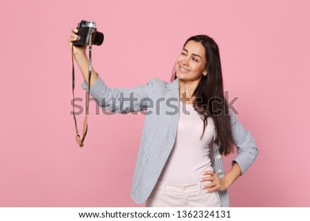 Portrait of blinking young woman in striped jacket doing selfie shot on retro vintage photo camera isolated on pink pastel background. People sincere emotions, lifestyle concept. Mock up copy space