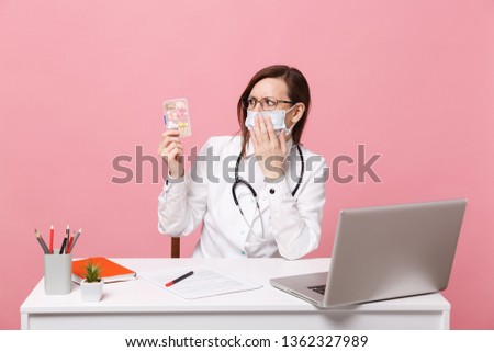 Female doctor sit at desk work on computer with medical document hold pills in hospital isolated on pastel pink wall background. Woman in medical gown glasses stethoscope. Healthcare medicine concept