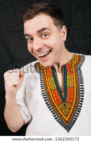 young cheerful guy in a national African shirt - dashiki. emotional portrait of a student. smiling male animator posing on a black background. clothes for party or fashion show