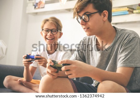 Young brothers enjoy video game at home. Smiling kids playing with console sitting on a sofa Teenager and brother playing with wireless remote controller Wifi technology is everywhere Friends have fun Royalty-Free Stock Photo #1362374942