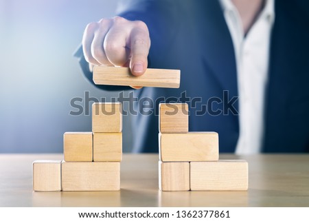 Business man bridging the gap between two towers or parties made from wooden blocks; conflict management or mediator concept, blue toned with ligth flare Royalty-Free Stock Photo #1362377861