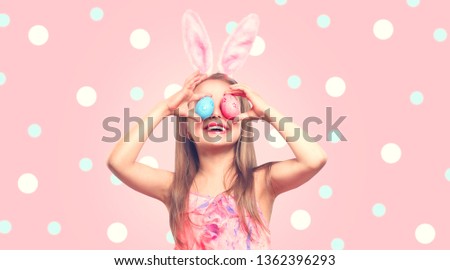 Funny Easter smiling little girl wearing bunny rabbit ears, holding colorful painted Easter eggs on her eyes. Baby girl laughing portrait over Polka Dots pink background. Joy. Spring holidays backdrop