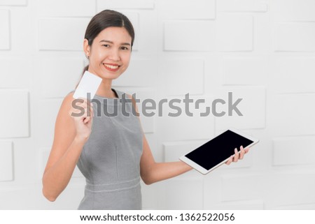 Half-length studio portrait of young beautiful, Asian lady, in gray sleeveless dress, holding tablet and white card, posing in advertising or online shopping concept, with white brick wall background