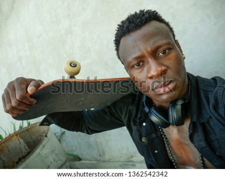 cool isolated portrait of young attractive and confident hipster black African American man leaning on street wall holding skate board as badass guy in urban style concept