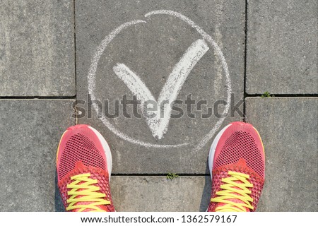 Checkmark ok sign on gray sidewalk with woman legs in sneakers, top view.
