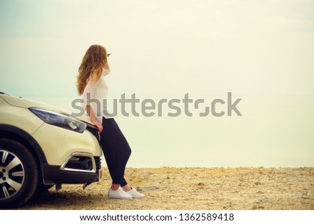 Happy stylish young woman traveler on beach road sitting on white crossover car, holding hat in hand. Banner. Travel, summer vacation, holiday, freedom concept. Digital detox.