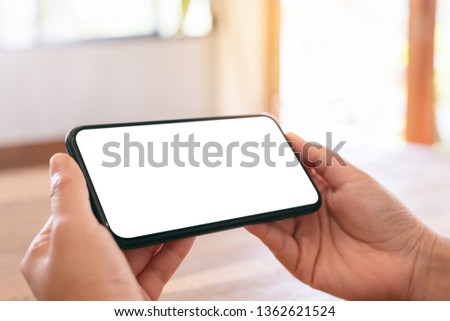 Mockup image of woman's hands holding black mobile phone with blank white screen horizontally on wooden table