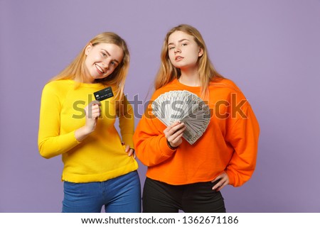 Two smiling blonde twins sisters girls holding credit bank card, fan of money in dollar banknotes, cash money isolated on violet blue background. People family lifestyle concept. Mock up copy space