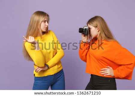 Two stunning young blonde twins sisters girls in colorful clothes taking pictures on retro vintage photo camera isolated on violet blue background. People family lifestyle concept. Mock up copy space
