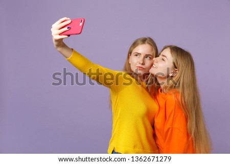 Two charming young blonde twins sisters girls in colorful clothes doing selfie shot on mobile phone isolated on pastel violet blue wall background. People family lifestyle concept. Mock up copy space
