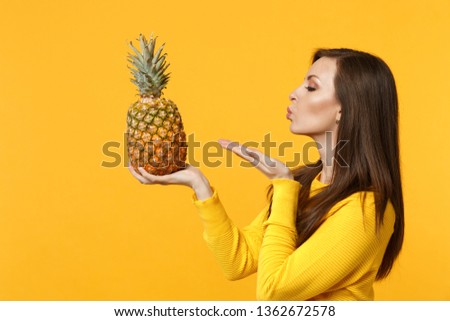 Side view of young woman in casual clothes sending air kiss holding fresh ripe pineapple fruit isolated on yellow orange background. People vivid lifestyle, relax vacation concept. Mock up copy space