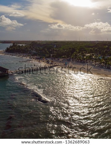 An aerial photo of the sun near sunset casting it's reflection in the water. The waves crash onto the beach as the pier is in the distance. 