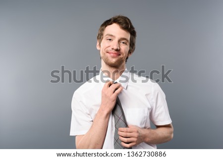 smiling businessman tying tie and looking at camera on grey background