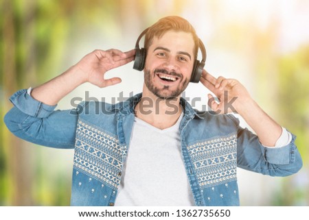 Smiling young man in headphones and casual clothes enjoying listening to music standing over blurred park background. Toned image
