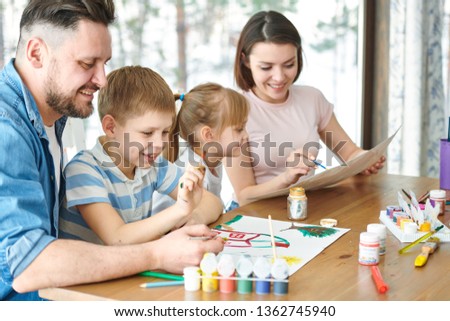 Young father and his son painting picture on paper with gouache of various colors while little girl and mother drawing with crayons near by