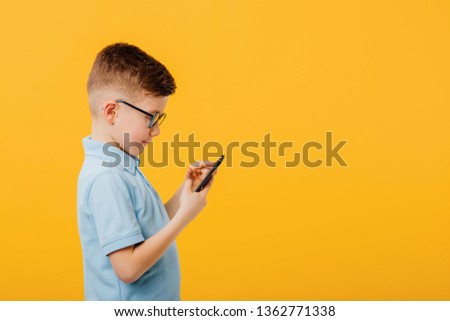 handsome little boy is concentrated on the phone, look at the smartphone, technology concept for children, in glasses, profile view,  dressed in blue shirt, isolated on yellow background, copy space Royalty-Free Stock Photo #1362771338