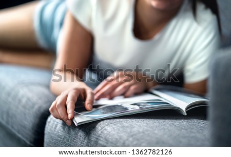 Woman lying on couch and reading a fashion or beauty magazine. Lady enjoying her day off with celebrity entertainment news or shopping catalogue. Hipster girl relaxing on sunday morning on comfy sofa. Royalty-Free Stock Photo #1362782126