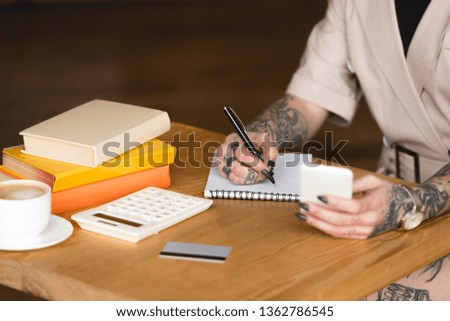 partial view of tattooed businesswoman writing in notebook and holding smartphone