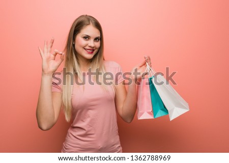 Young pretty russian girl cheerful and confident doing ok gesture. She is holding a shopping bags.