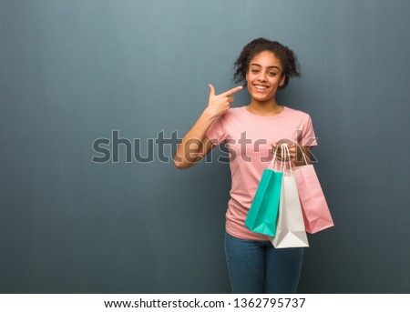 Young black woman smiles, pointing mouth. She is holding a shopping bags.