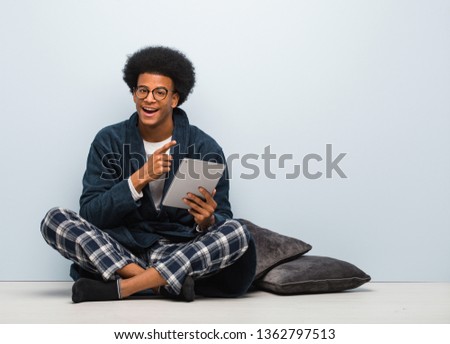 Young black man sitting on his house and holding his tablet pointing to the side with finger
