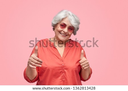 Beautiful elderly female in trendy outfit and sunglasses cheerfully smiling and showing thumb up gesture with both hands while standing against pink background. Hipster grandmother