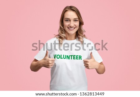 Pretty young woman in white T-shirt with volunteer writing smiling and showing thumb up gesture with both hands while standing on pink background