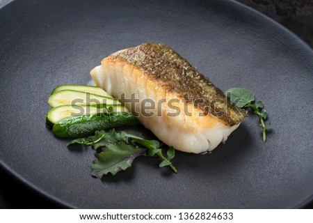 Fried Danish skrei cod fish filet with baby zucchini and lettuce as closeup on a modern design plate 