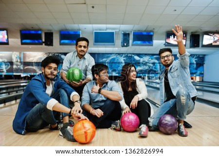 Group of five south asian peoples having rest and fun at bowling club, sitting on bowling alley with balls on hands.