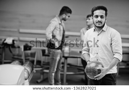 South asian man in jeans shirt standing at bowling alley with ball on hands. Guy is preparing for a throw.