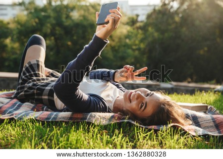 Cheerful teenage girl laying on a blanket at the park, relaxing, taking a selfie