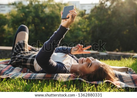 Cheerful teenage girl laying on a blanket at the park, relaxing, taking a selfie