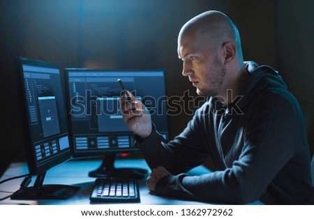 cybercrime, hacking and technology concept - male hacker with smartphone and progress loading bar on computer's screens in dark room Royalty-Free Stock Photo #1362972962