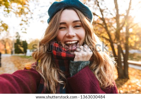 Cheerful young girl with long brown hair wearing autumn coat, walking at the park, taking a selfie