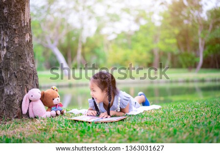 cute little girl  reading a book while lying with a doll in the park