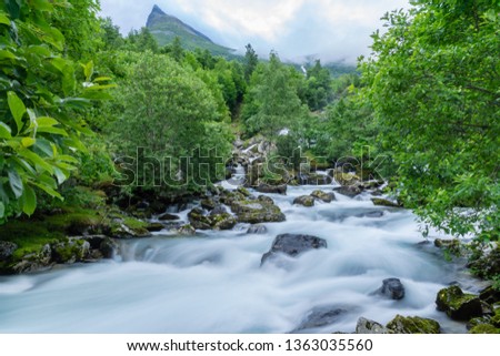 Landscape from the amazing Norwegian mountains,  a creek with flushing white soft water streaming down a mountainside with lush green trees and a mountain peak in the background