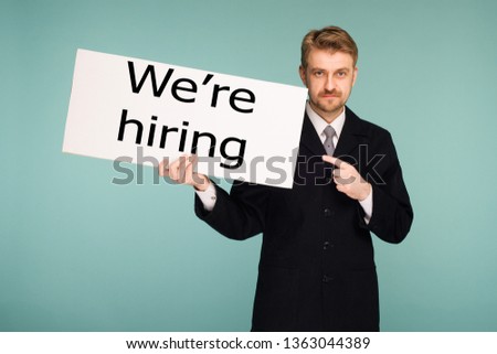 Happy smiling young business man points finger signboard with sing we're hiring, on blue background