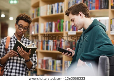 Serious young multiethnic student boys in casual clothing standing in library and choosing literature: black guy reading book, young man in hoodie using mobile app on smartphone