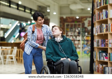 Positive attractive young woman with satchel pushing wheelchair and talking to disabled student in library Royalty-Free Stock Photo #1363058348